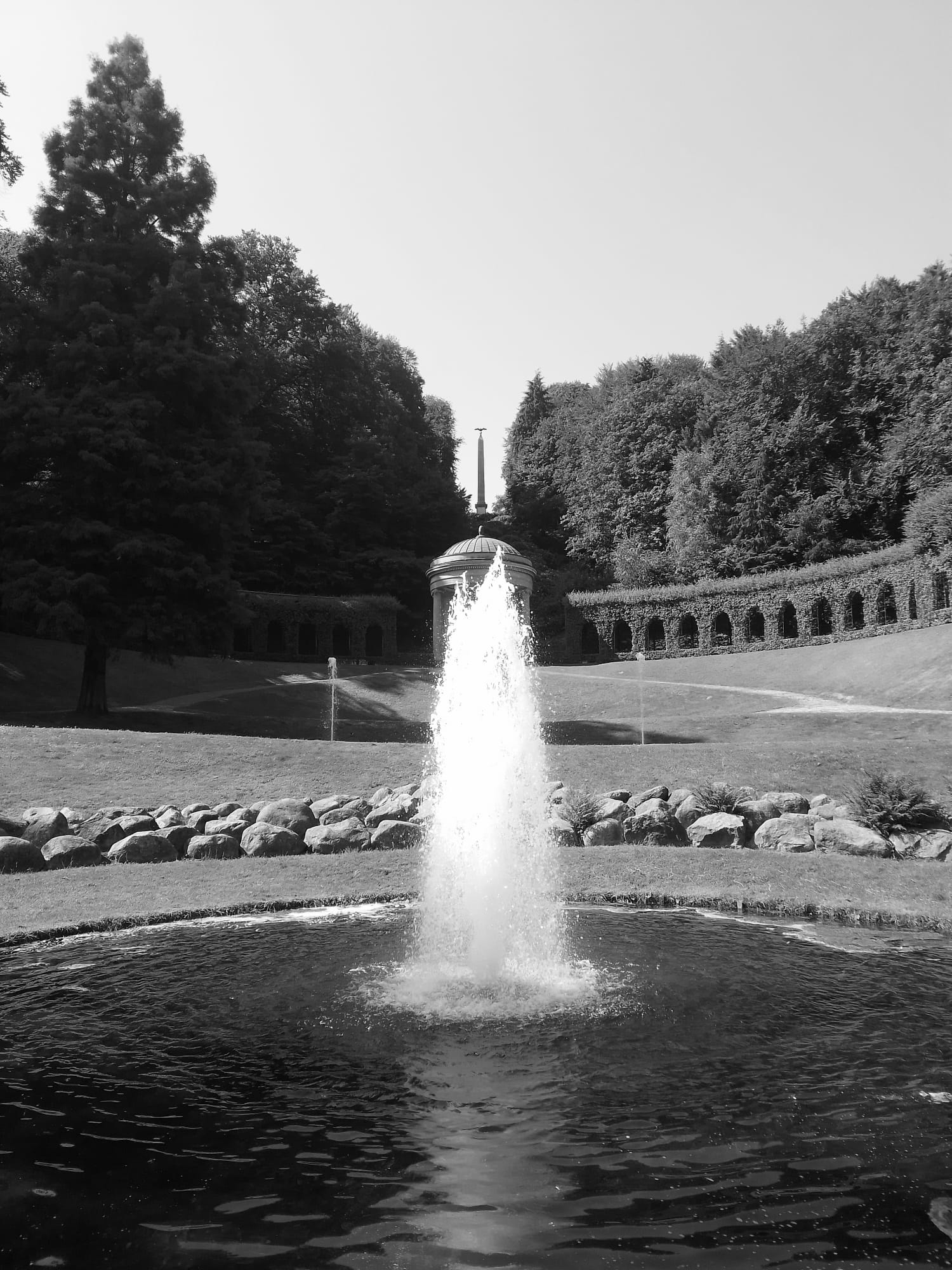 A Black and white photo of a fountain with a domed pavilion in the background and an obelisk type structure behind it.