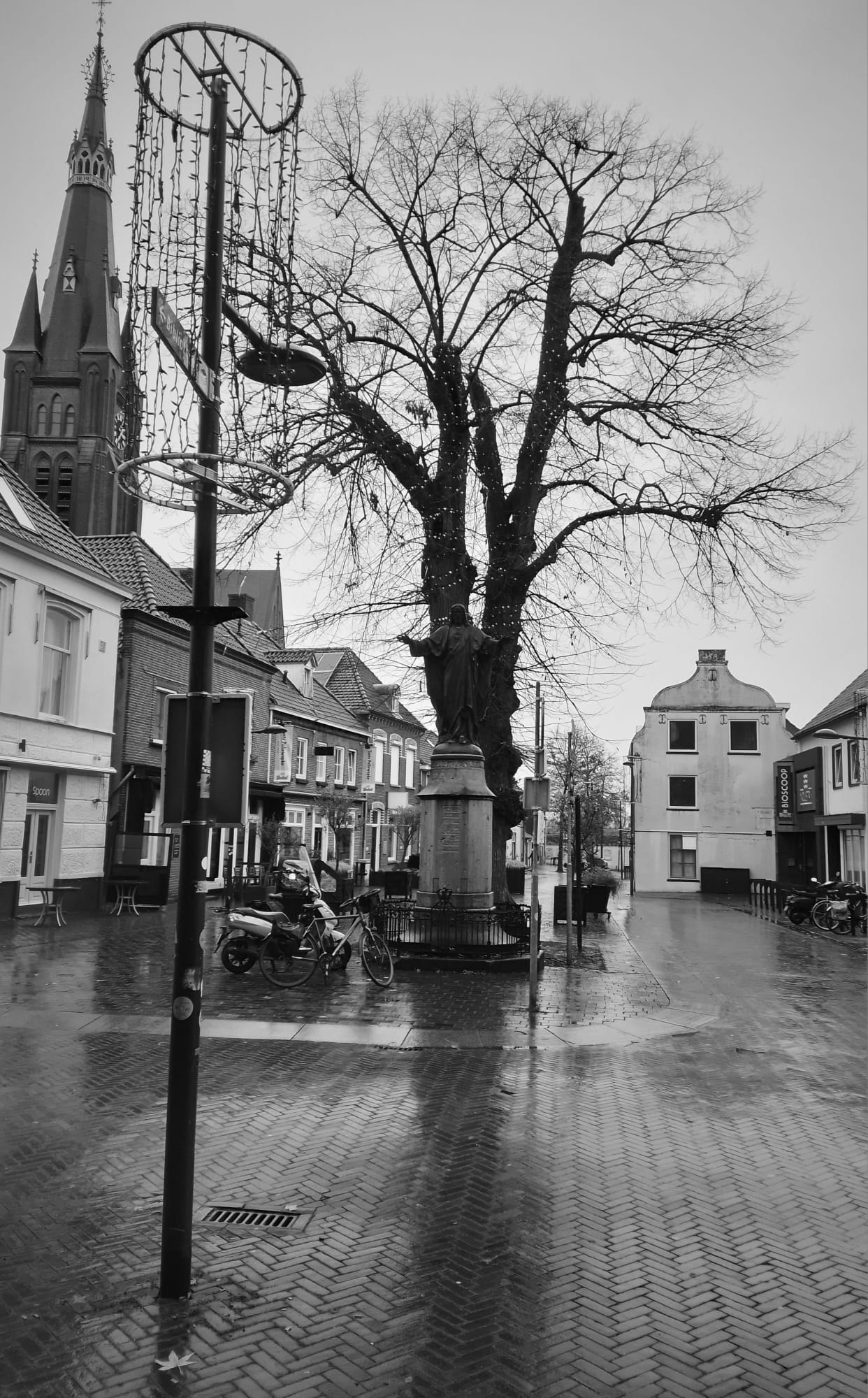 A Black and white picture looking at a statue of Jesus, a tree directly behind it, a few other buildings and a church.